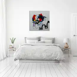 Tableau style pop art d'un éléphant multicolore, accroché au-dessus d'un lit à la parure grise et blanche, 2 tables de chevet avec sur la gauche une plante verte , un réveil et à droite une lampe