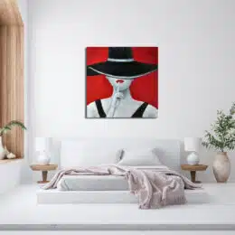 Tableau rouge d'une femme vêtu d'un grand chapeau noir, son index sur sa bouche rouge, accroché au-dessus d'un lit avec 2 tables de chevet en bois et 2 lampes blanches, un plaid rose déborde sur le bout du lit et un vase avec une plante est sur la droite à côté de la table de chevet