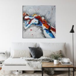 Tableau d'une silhouette féminine sur fond gris, de couleur bleu et rouge sur le corps, le bras droit sous la tête accroché au-dessus d'un canapé large avec 2 gros coussins blanc, un petit noir et 2 petits beiges, une table basse carrée en bois et support en fer noir avec une plante verte une grande lampe noire posé sur la droite du canapé