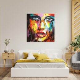 Tableau style popart d'un visage d'une femme aux couleurs vivent, regard aux yeux bleus, accroché au-dessus d'un lit à la parure blanc et jaune avec 2 tables de chevet en bois à gauche un vase avec une fleur, un réveil et un fauteuil gris sur la droite