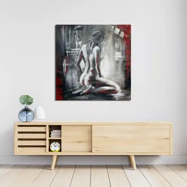 Tableau noir et blanc d'une femme nue à genoux, assise sur ses talons de dos, les cheveux mi-long sur fond gris, noir avec les côtés rouges, accroché au-dessus d'un meuble en bois clair à porte coulissante avec un vase posé sur la gauche