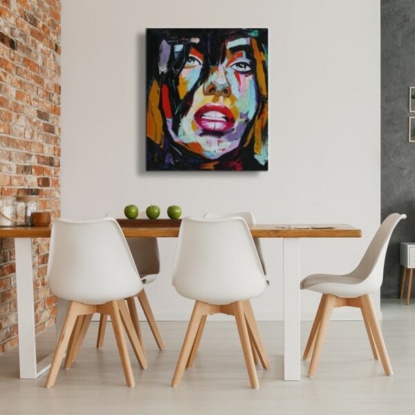Tableau d'un visage d'une femme style pop art, accroché en face d'une table en bois contre un mur en pierre avec 5 chaises blanche avec les pieds en bois