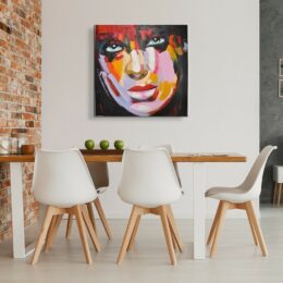 Tableau style pop art d'un visage d'une femme, accroché face à une table en bois avec 5 chaises blanches avec les pieds en bois