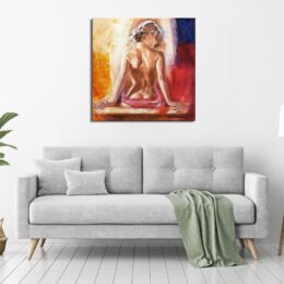 Tableau avec des tons chauds, rouge et jaune, d'une femme de dos nue, les cheveux attachés, les fesses posées contre une table et les mains appuyées de chaque côté, accroché au-dessus d'un canapé gris avec un plaid tombant par terre