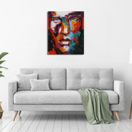 Tableau d'un visage en gros plan aux couleurs vives, accroché au-dessus d'un canapé gris avec un plaid vert tombant