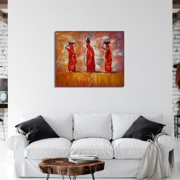 Tableau de 3 femmes africaines portant des corbeilles sur la tête dans un décor désert aux tons oranges, rouges , accroché au-dessus d'un canapé blanc avec des coussins noirs de chaque côté et une table basse en bois marron