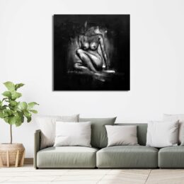 Tableau noir et blanc d'une femme assise nue les jambes pliées sur le côté et les mains posées sur le côté de ses hanches, au-dessus d'un canapé kaki, des coussins blanc, beige, avec une plante verte dans un pot en terre sur la gauche